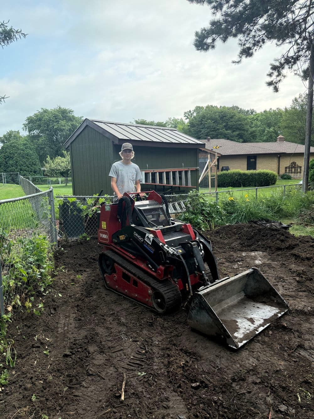 Young man operating a compact track loader in a garden area with a home in the background.